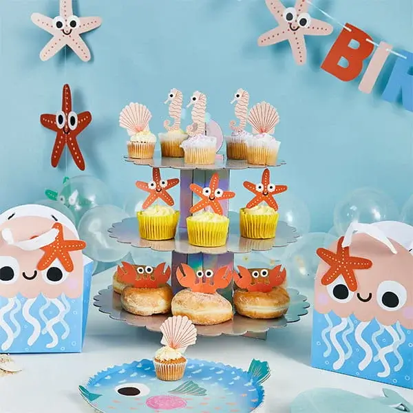 Under-the-sea themed cupcakes and doughnuts displayed on a tiered stand with starfish, seahorse, and shell decorations, surrounded by ocean-themed party tableware.